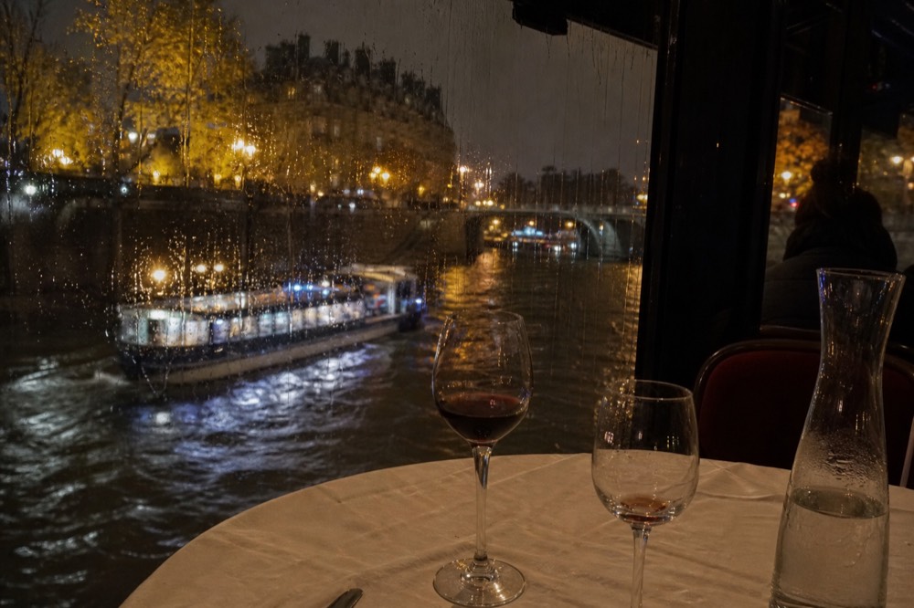Set dinner table with wine glasses beside a window overlooking the Seine at night, no text, no logos, no people.