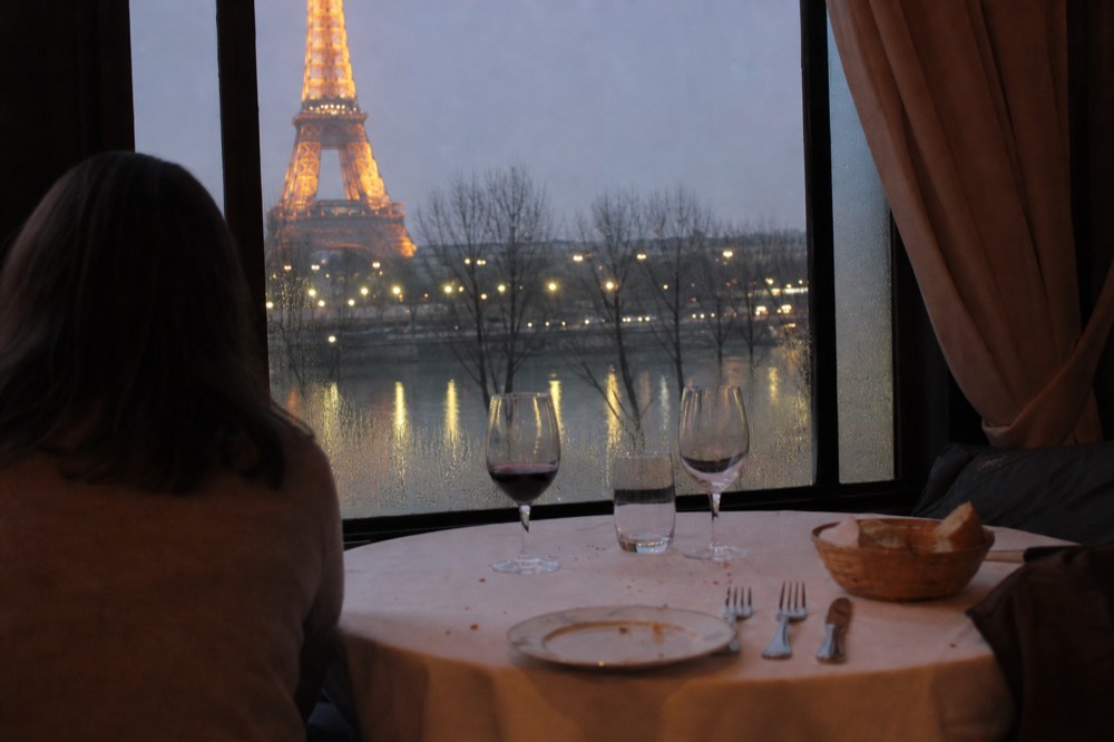 Couple's place setting on a dinner cruise with the illuminated Eiffel Tower reflected in the river outside the window, no text, no logos, no people.