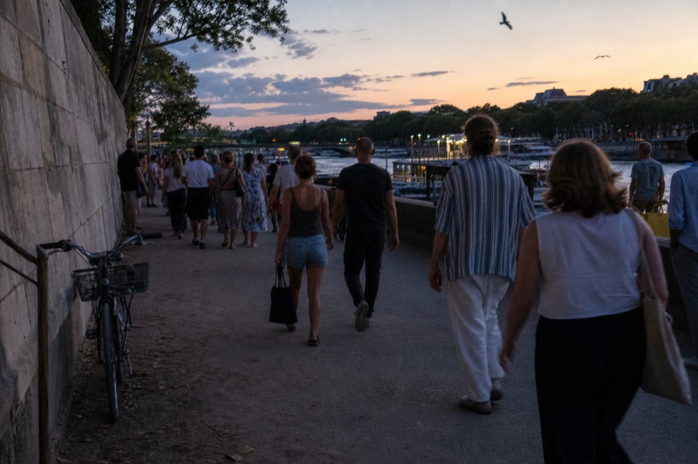 Evening crowd walking along the Seine quays toward moored boats, no text, no logos, no people
