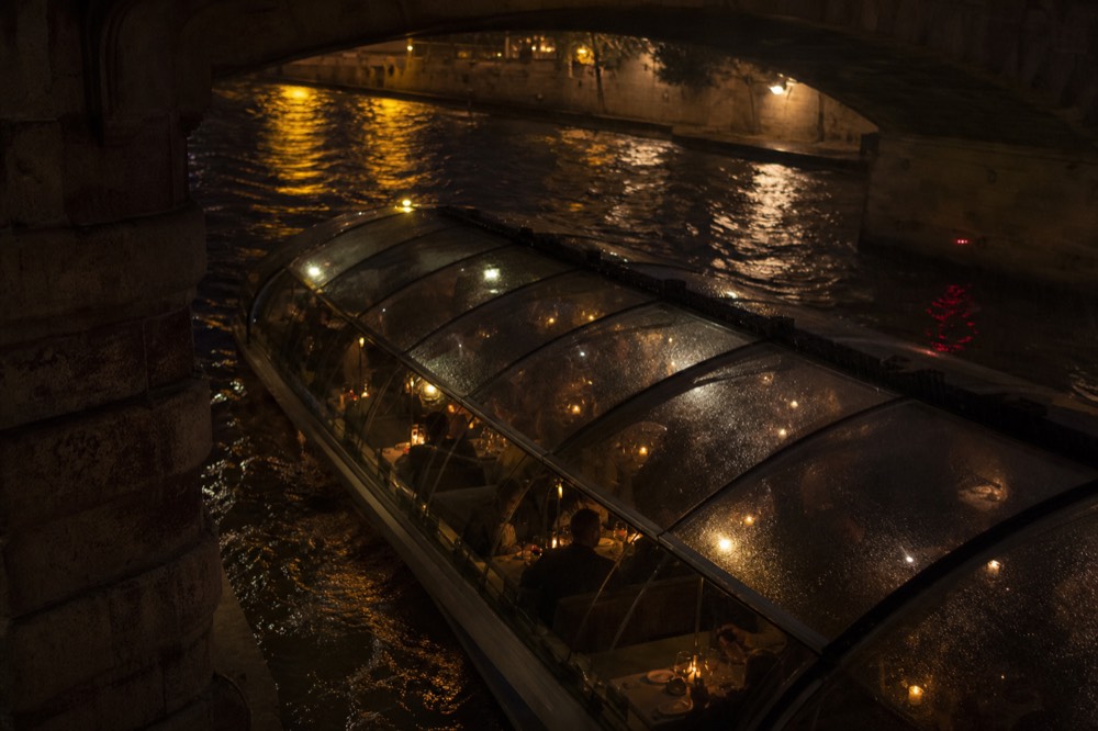 Glass-roofed dinner boat passing under an illuminated Paris bridge at night, no text, no logos, no people